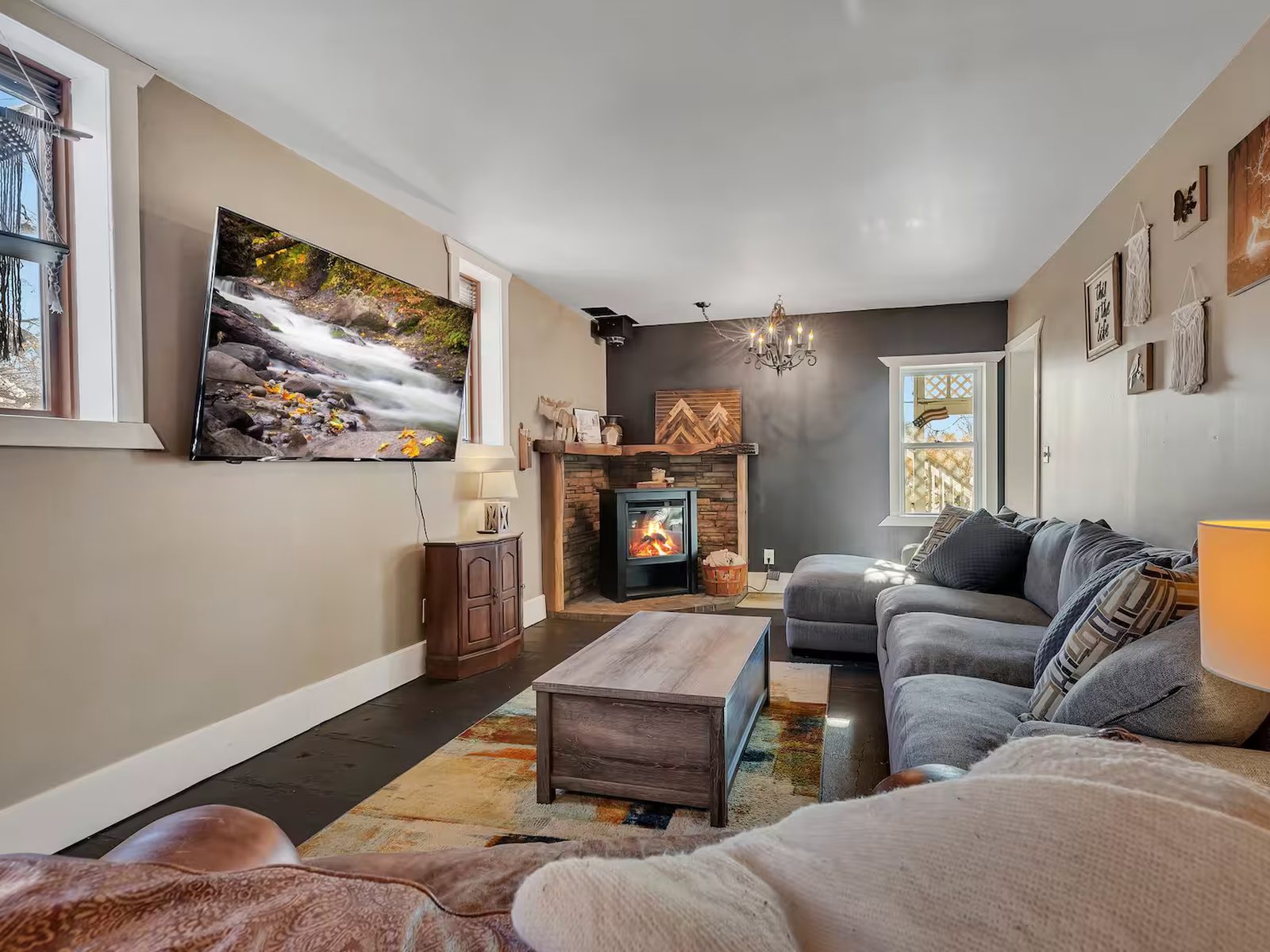 The living room at Sunnyside Manor: a wood stove set into stone beneath a live-edge mantel, a grey sectional, dark floors and a chandelier.